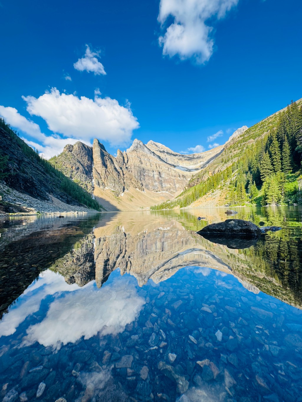 A crystal-clear alpine lake reflects jagged mountain peaks, a bright blue sky, and fluffy white clouds. The lake's shallow, rocky bottom is visible beneath the reflection, creating a mirror-like effect. Surrounded by evergreen trees and dramatic cliffs, the serene landscape captures the natural beauty of a high-altitude wilderness, in Banff National Park.